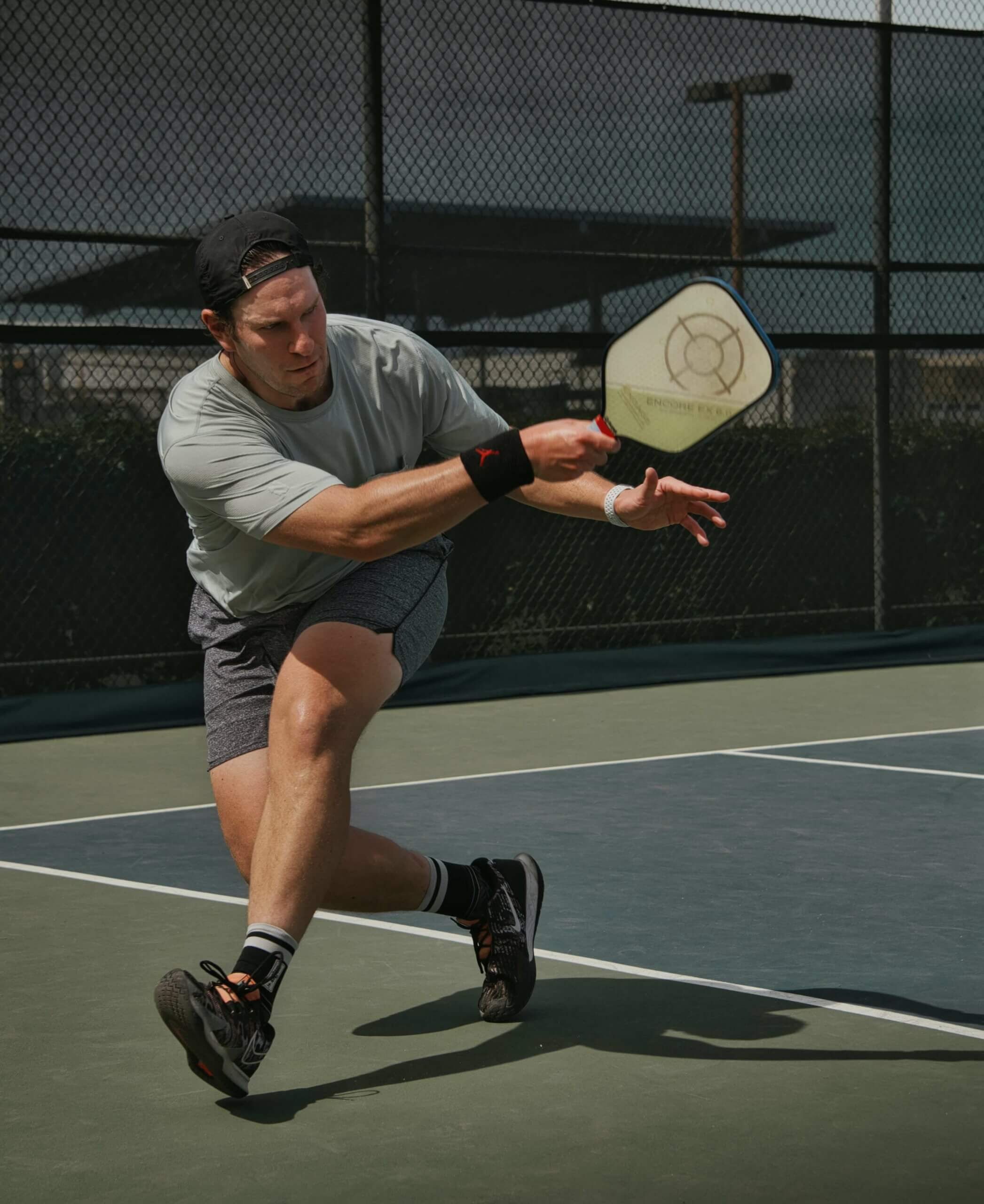 A man playing pickleball on an outdoor court, swinging his paddle to hit the ball near a chain-link fence